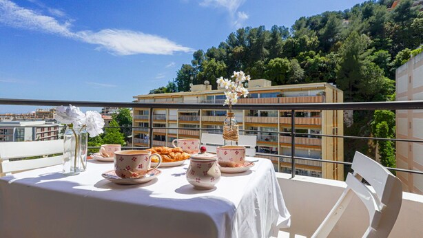 Un balcon avec une table dressée pour le thé, entouré de chaises blanches, d’où l’on peut admirer un panorama urbain avec ses bâtiments et ses arbres.