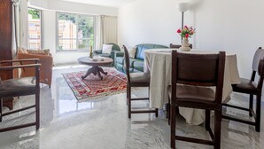 Un salon bien éclairé, doté d’un sol en marbre, d’un canapé vert, d’une table à manger avec des chaises et offrant une vue dégagée grâce à de grandes fenêtres.