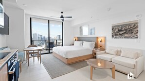 A modern hotel room with a large bed, a sofa, a dining table, and a city view.