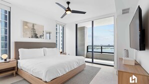 A modern bedroom with a large bed, a ceiling fan, a TV, and a balcony with a city view. - Stunning Ocean Views, Prime Pool & Gym Access (Miami)