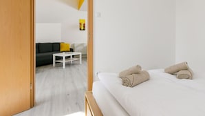 A modern hotel room with a wooden door, a bed with rolled towels, a white bedside table, and a grey sofa with a yellow pillow in the background.
