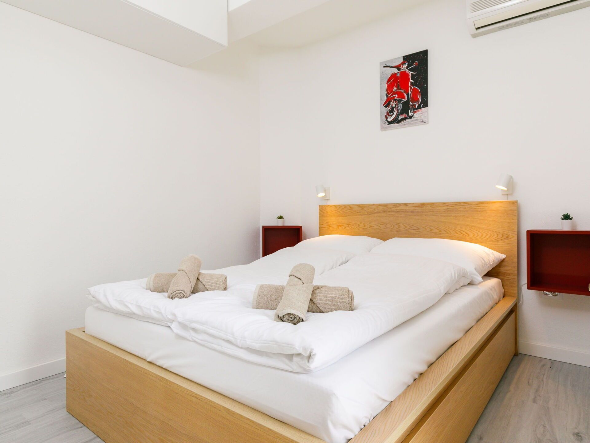 Een netjes opgemaakt bed met witte lakens en opgerolde handdoeken, een houten hoofdbord en een klein schilderij van een rode scooter aan de muur.