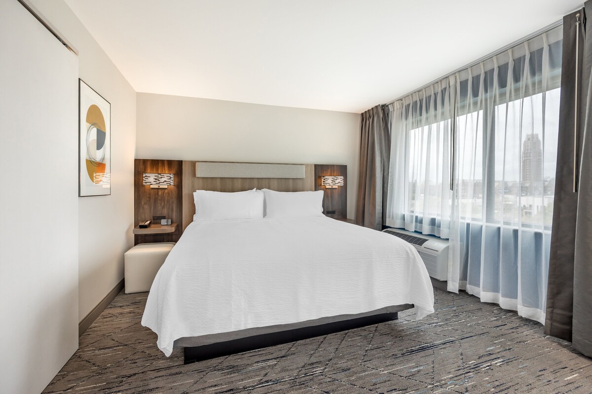 Habitación de hotel con una cama grande, una mesita de noche, aire acondicionado de pared y vistas a la ciudad a través de cortinas translúcidas.