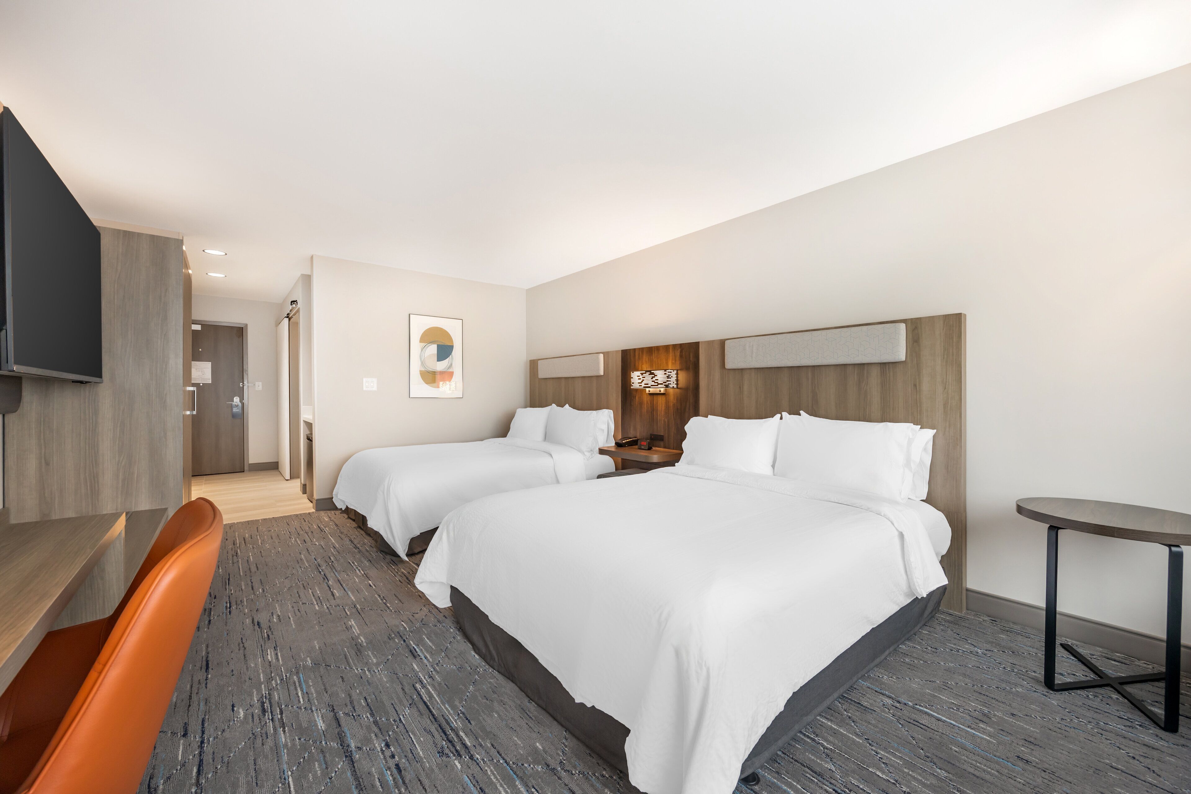 A hotel room with two beds, a desk, a chair, a small table, and a TV.
