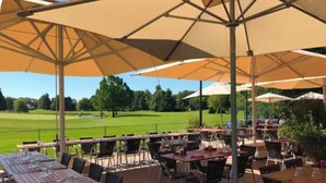 Ein Außenbereich zum Essen mit Tischen, Stühlen und großen Sonnenschirmen, mit Blick auf einen Golfplatz.
