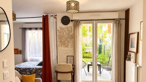 Ein modernes Zimmer mit einem Bett, einem Sofa, einem Esstisch und Blick auf einen Garten durch eine Schiebetür.