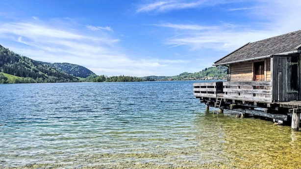 Eine Holzhütte auf Stelzen über einem klaren See, im Hintergrund ein bewaldeter Berg.