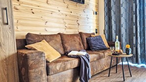 Ein Zimmer mit Holzwänden, einem braunen Ledersofa, einem kleinen runden Tisch mit einer Flasche, Gläsern und Snacks sowie einem Fenster mit gemusterten Vorhängen.