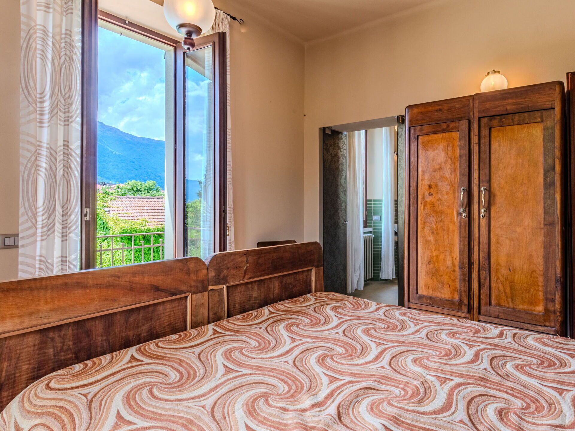 A bedroom with a large bed, a wooden wardrobe, and a window with a view of greenery and mountains.
