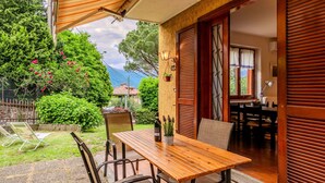 An outdoor patio with a wooden table and chairs, a covered seating area, and a view of a house and greenery in the background. - Lovely apartment for 4 people with WIFI, A/C, TV, patio and pets allowed (Bellano)