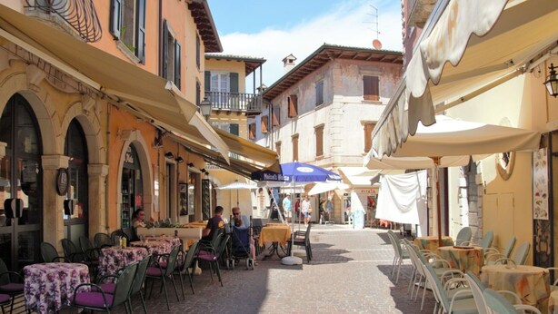 A cobblestone street with outdoor seating and umbrellas, surrounded by historic buildings with balconies and arches. - Nice apartment for 8 people with pool and terrace (Torri Del Benaco)