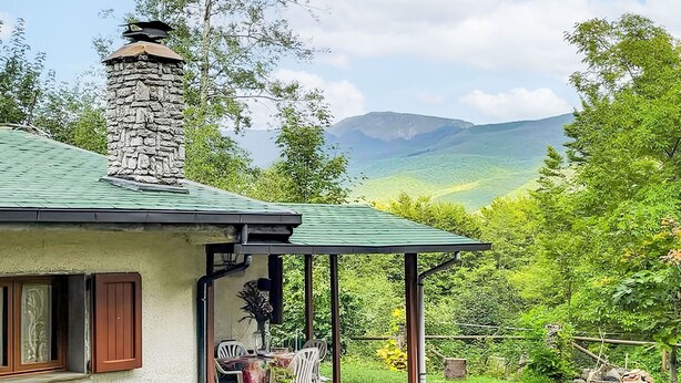 Een stenen huis met een groen dak, schoorsteen en terras met een tafel en stoelen.