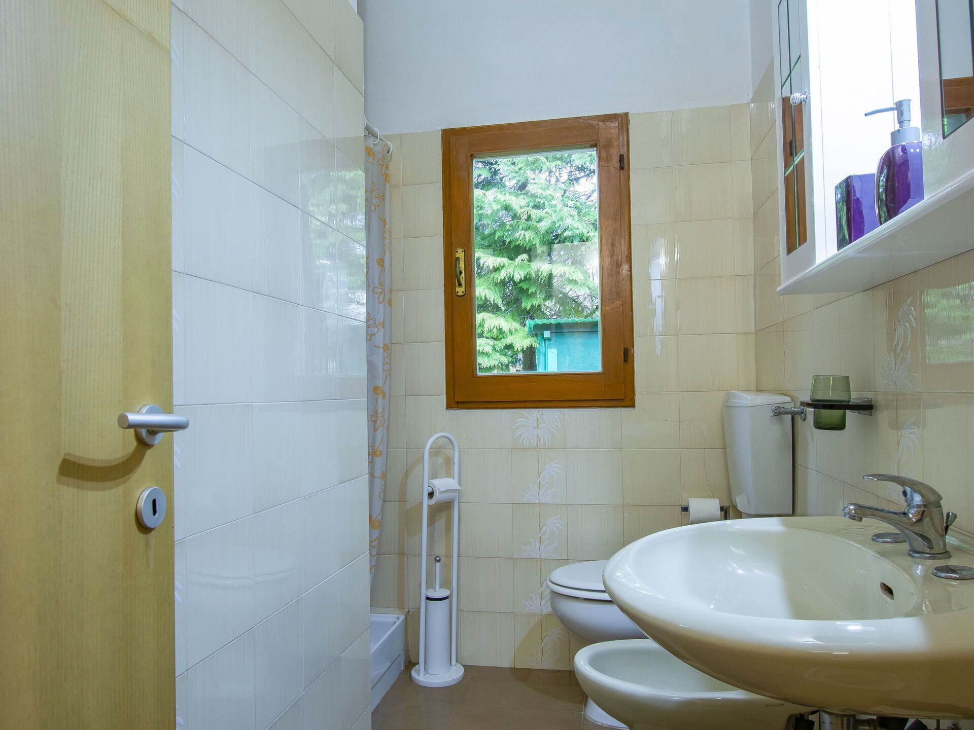 Ein Badezimmer mit Toilette, Bidet und Waschbecken. Ein Fenster bietet Blick auf Grünflächen außerhalb.