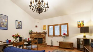 Een woonkamer met een open haard, blauwe banken, een eettafel en een kroonluchter.