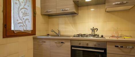 A modern kitchen with wooden cabinets, a gas stove, an oven, and a window with a lace curtain.