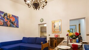 Een woonkamer met een blauwe bank, een eettafel met borden en bestek, een kroonluchter en een schilderij aan de muur.