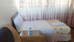 Ein Zimmer mit braunem Sofa, einem Bett mit gemusterten Betttextilien, einem kleinen Tisch mit einer Wasserflasche und einem Vorhang mit Karomuster.