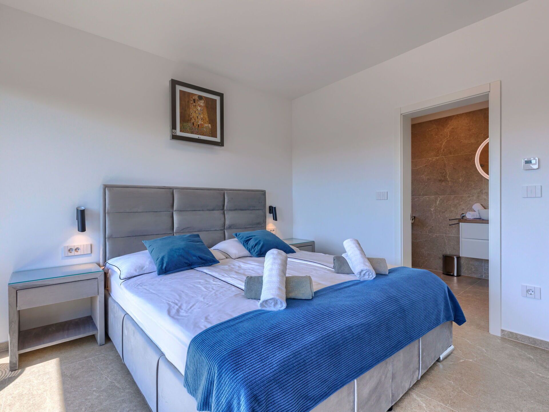 Ein modernes Schlafzimmer mit einem großen Bett, einem Nachttisch, einer Wandlampe und einem gerahmten Bild an der Wand.