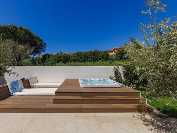 A wooden deck with a hot tub, a seating area with cushions, and a view of a hillside with trees and buildings.