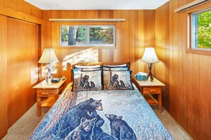 A bedroom with wooden walls, a bed with a bear-themed duvet, two bedside tables with lamps, and a window with a view of trees.