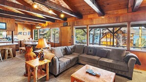Interior acolhedor de uma cabana, com teto de madeira, uma janela ampla com vista para outra cabana e árvores, um sofá, uma mesa de centro e um abajur.