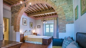 Un dormitorio rústico con paredes de ladrillo visto, techo de madera, cama con ropa de cama floral, mesita de noche de madera, un banco y una puerta con ventanal.