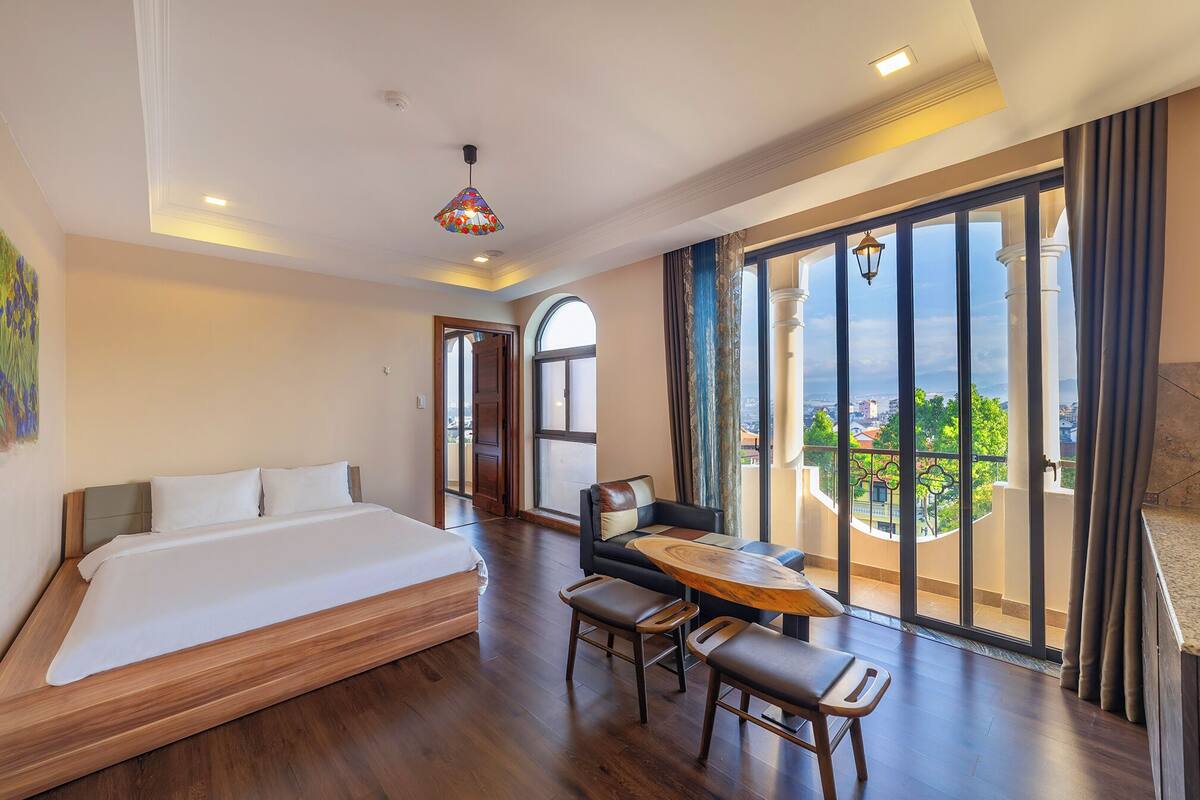 Habitación de hotel con cama, una mesa de comedor con sillas, balcón con vistas y una lámpara decorativa colgante.