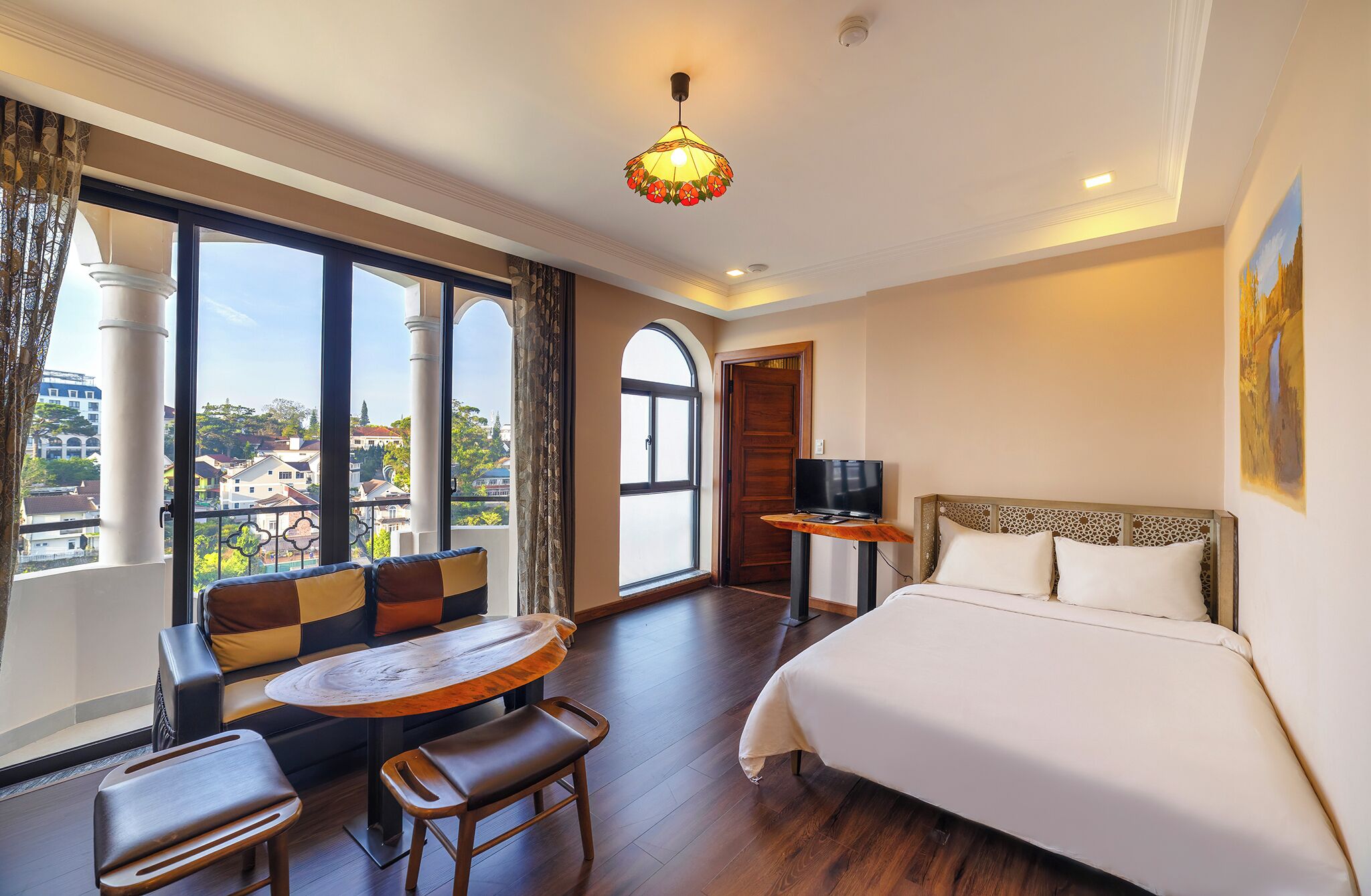 Ein Hotelzimmer mit einem großen Bett, einem Fernseher auf einem Holzständer, einem runden Holztisch mit Stühlen und Blick auf die Stadt durch große Fenster.