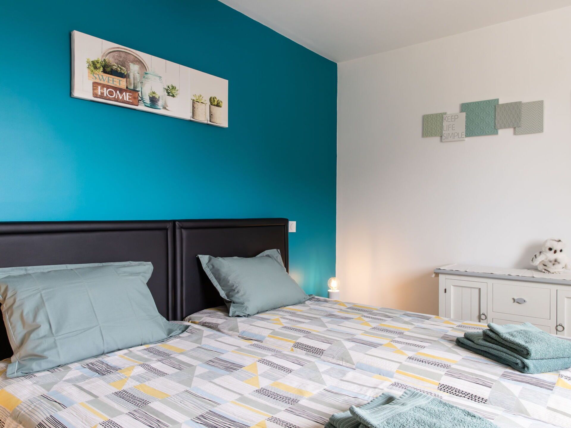 Een slaapkamer met een bed, kussens, een nachtkastje, een muur met een bord en een kast.