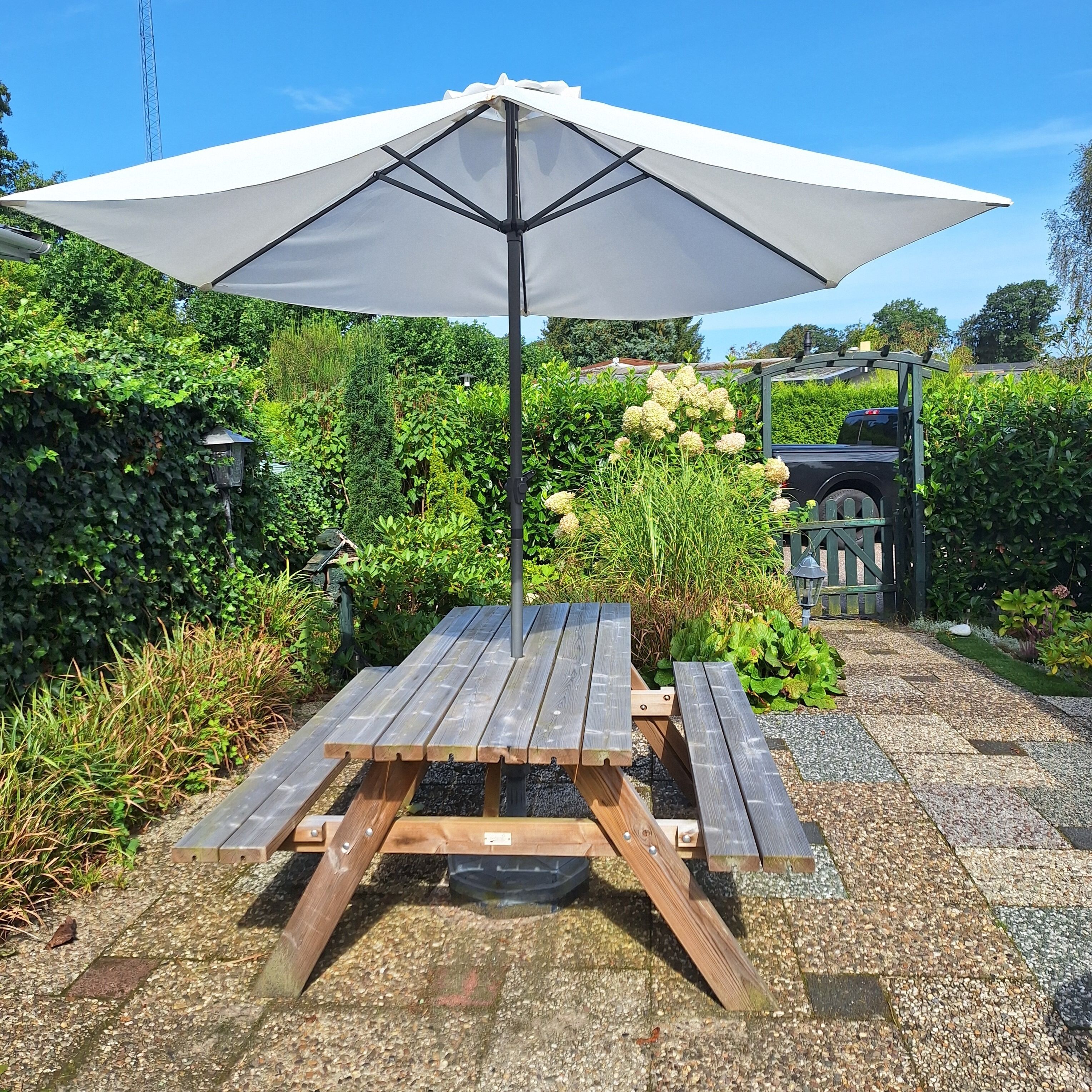 A wooden picnic table with a white umbrella, surrounded by lush greenery and a gravel path.