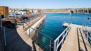 Une terrasse en bois avec des chaises longues et un espace aménagé au bord de la piscine, d’où l’on peut admirer une étendue d’eau calme, des bateaux et une côte rocheuse en arrière-plan.
