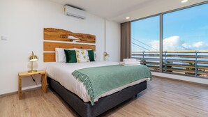 Une chambre moderne avec un grand lit, une tête de lit en bois et une vue sur la ville grâce à de grandes fenêtres du sol au plafond.