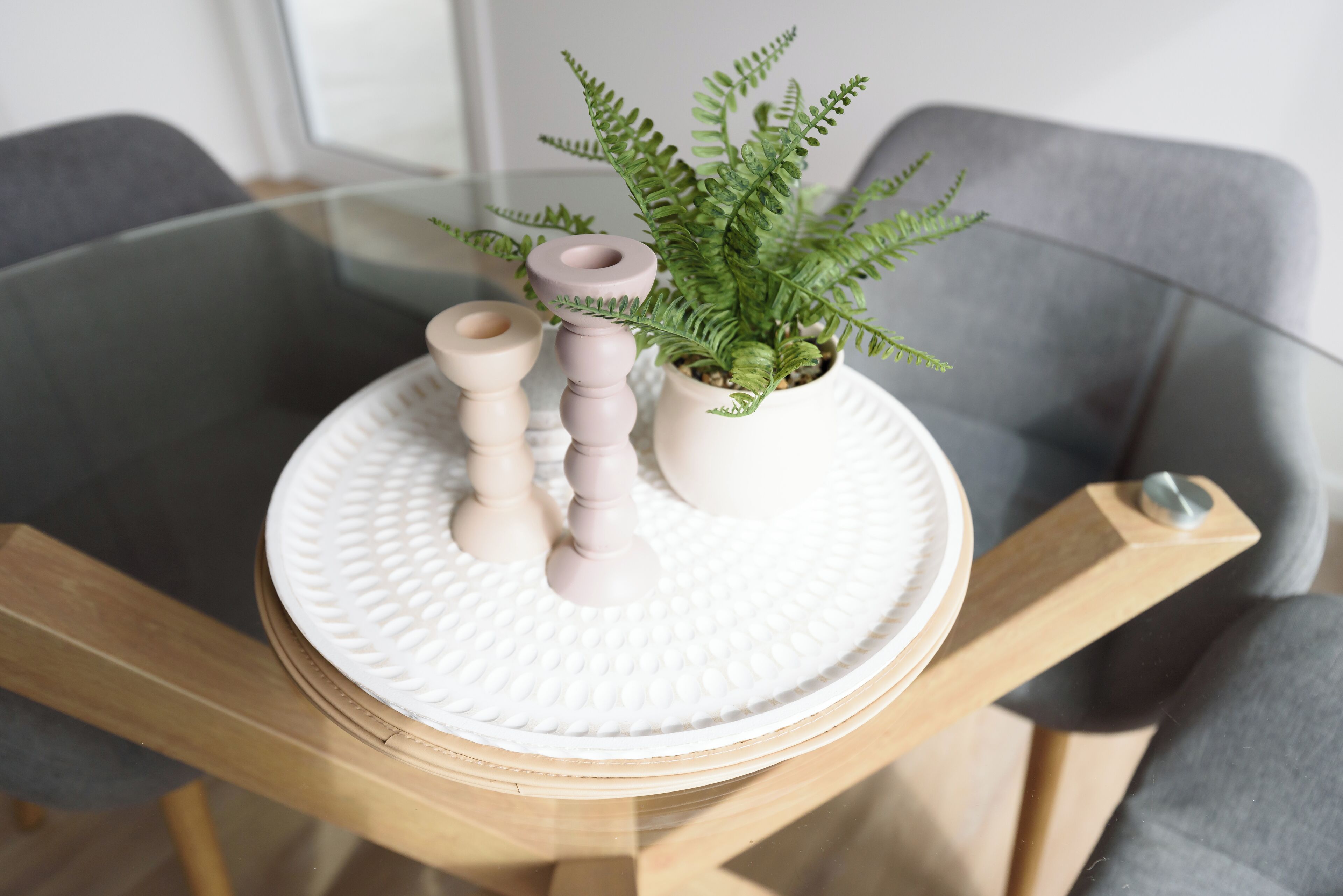 Una mesa con cubierta de vidrio, una bandeja blanca con textura, una pequeña planta en maceta y dos portavelas de madera.