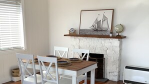 A dining area with a wooden table and white chairs, a fireplace with a sailboat painting above it, and a hanging light fixture.