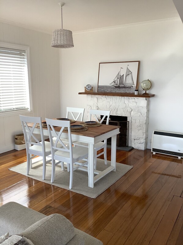A dining area with a wooden table and white chairs, a fireplace with a sailboat painting above it, and a hanging light fixture.