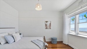 A bedroom with a large bed, a bedside table, a small stool, a framed picture, and a window with a view of the sea.