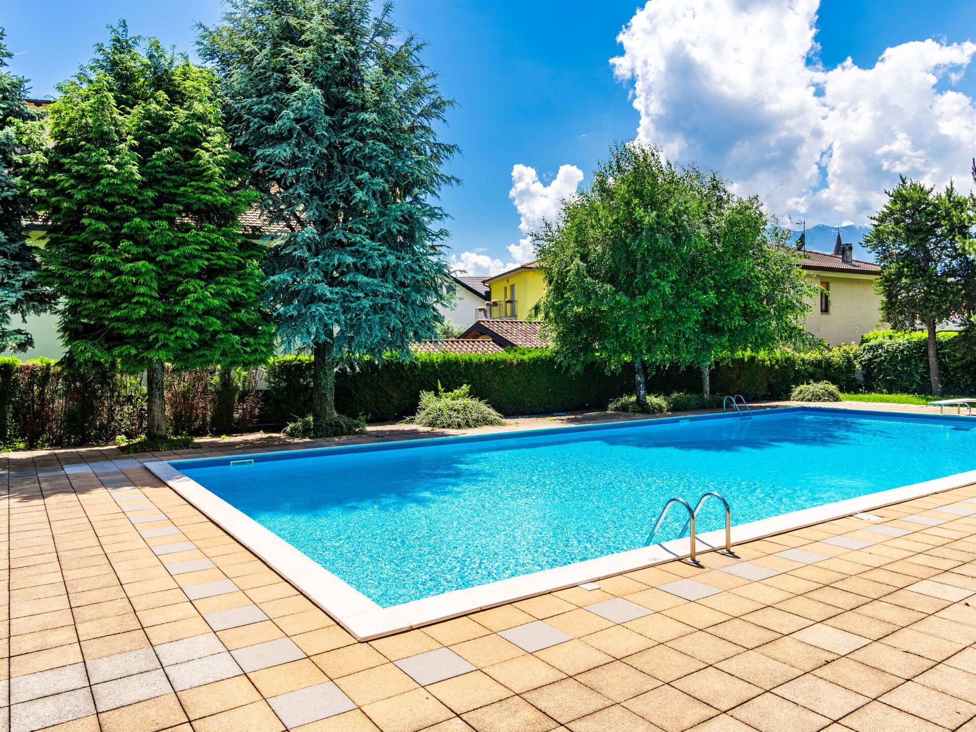 A rectangular swimming pool with a tiled deck, surrounded by lush greenery and a clear blue sky.