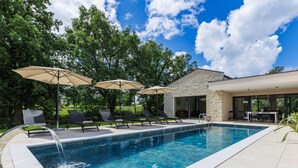 A rectangular swimming pool with a waterfall feature, surrounded by lounge chairs and umbrellas, with a stone building and a covered patio area in the background.