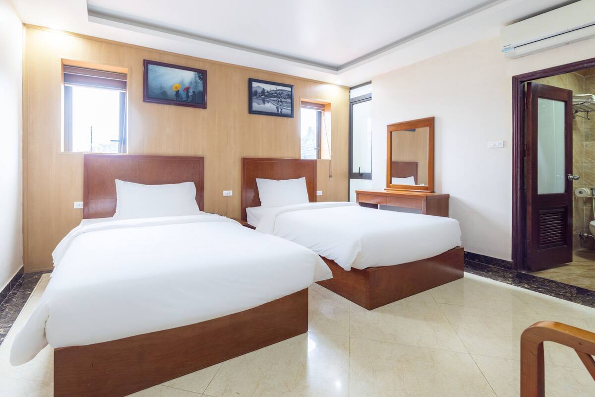 Quarto de hotel com duas camas, cabeceiras de madeira, cômoda de madeira, um espelho e banheiro com vaso sanitário e pia.