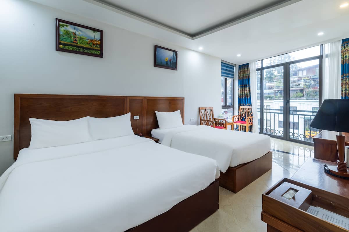 Quarto de hotel com duas camas, uma cabeceira de madeira, uma escrivaninha com abajur e janelas amplas que oferecem vista para o exterior.