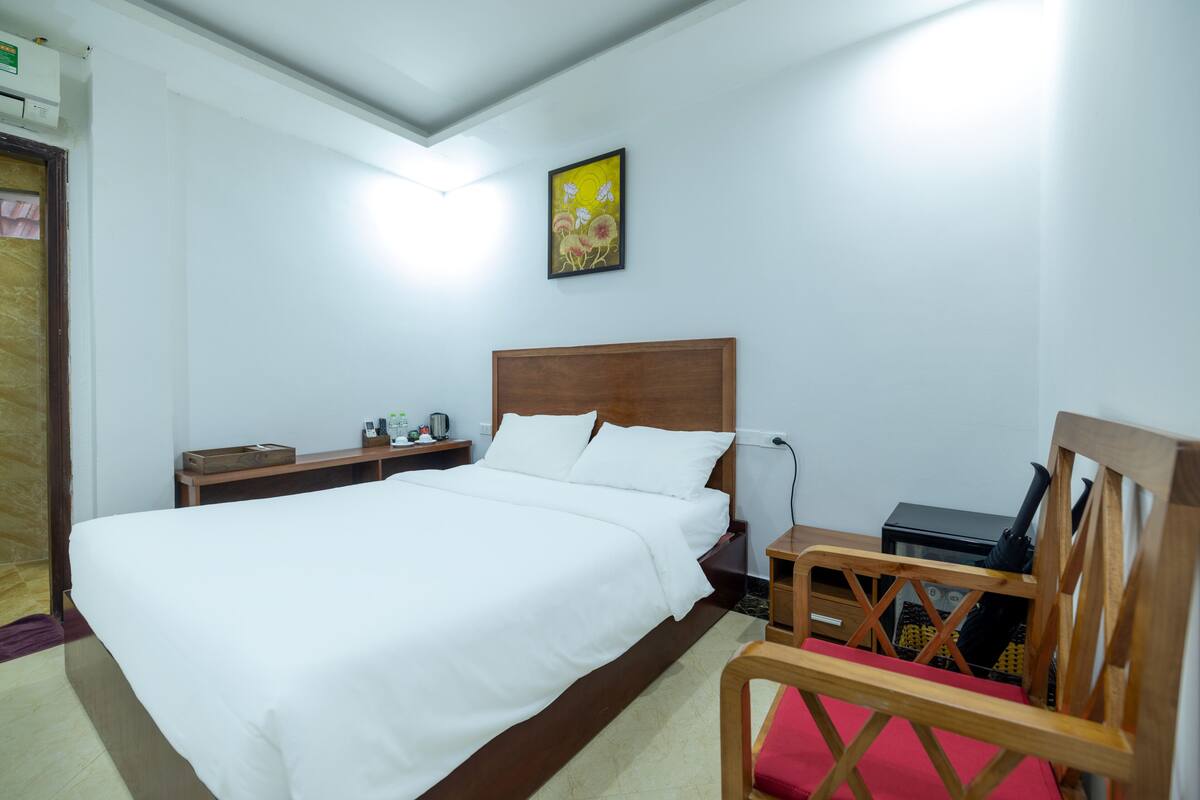 Uma cama bem arrumada com lençóis brancos, cabeceira de madeira, mesa de cabeceira com abajur e uma cadeira com almofada vermelha.