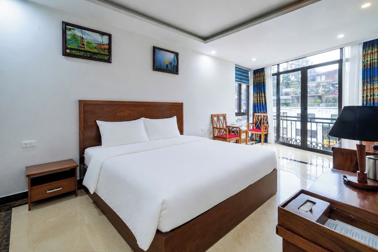 A hotel room with a large bed, a nightstand, a desk with a lamp, two chairs, and a window with a view of the city.
