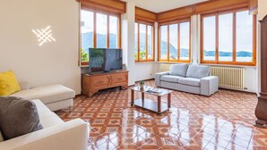 A living room with a TV, sofa, and coffee table, large windows with a mountain view, and patterned floor tiles. - Stunning private villa for 11 guests with WIFI, TV, balcony and pets allowed (Arizzano)