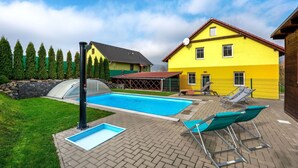 Ein Garten mit Swimmingpool, zwei Liegestühlen, einem Gewächshaus und einem gelben Haus.