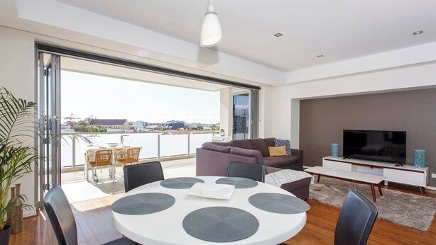 A modern living room with a round dining table, a sofa, a TV, and a balcony with a view of the city. - “Enjoy Fremantle from this spacious 3BR apartment with port views, communal pool & free parking — walk to Rottnest Ferry & Markets.” (Fremantle)