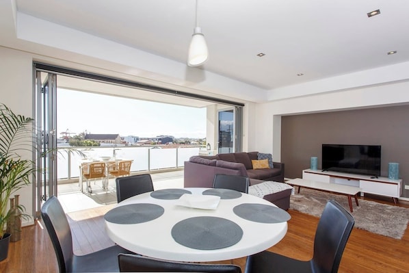 A modern living room with a round dining table, a sofa, a TV, and a balcony with a view of the city.