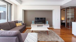 A modern living room with a flat-screen TV on a white stand, a grey sofa with cushions, a patterned throw, and a shaggy rug. - “Enjoy Fremantle from this spacious 3BR apartment with port views, communal pool & free parking — walk to Rottnest Ferry & Markets.” (Fremantle)