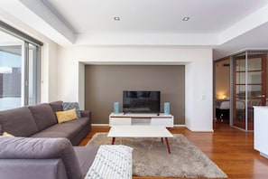 A modern living room with a flat-screen TV on a white stand, a grey sofa with cushions, a patterned throw, and a shaggy rug.