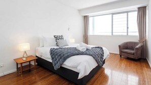 A modern bedroom with a large bed, bedside lamps, a chair, and a window with curtains. - “Enjoy Fremantle from this spacious 3BR apartment with port views, communal pool & free parking — walk to Rottnest Ferry & Markets.” (Fremantle)