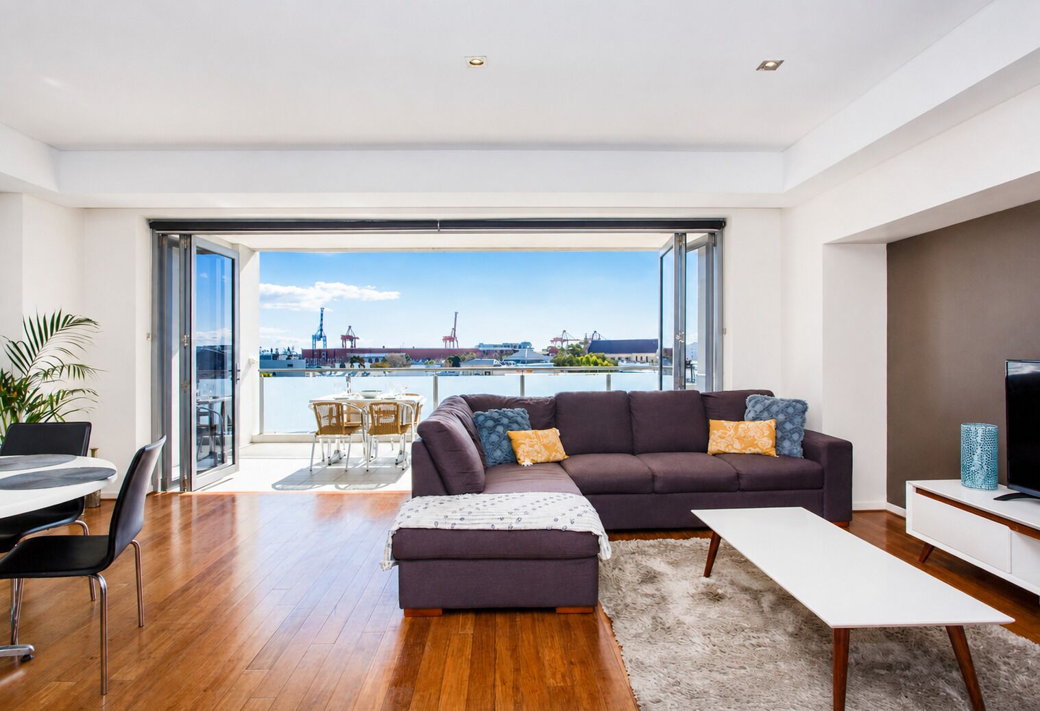 A modern living room with a sofa, a coffee table, and a TV stand. There is a balcony with a view of the city.